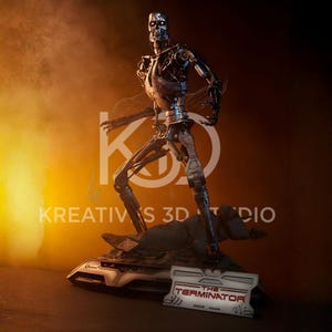 May include: A metallic silver robotic endoskeleton figure standing on a dark, textured base. The figure has glowing red eyes and exposed mechanical joints. A rectangular plaque in front displays the text THE TERMINATOR in red and silver.