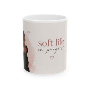 May include: A white ceramic mug featuring a graphic illustration of a person in a black jumper holding a cup. The text "soft life in progress" is printed in a mix of serif and script fonts, accompanied by two small outlined heart shapes.