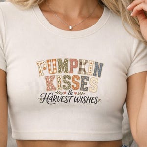 May include: A natural ivory ribbed short-sleeve crop top featuring the text PUMPKIN KISSES & HARVEST WISHES in a multi-colored patterned block font. The design includes small heart and leaf accents in brown, orange, and blue tones.