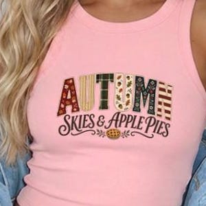 May include: A light pink ribbed tank top featuring the text "AUTUMN SKIES & APPLE PIES" in a multi-patterned font. The letters are filled with various autumn-themed prints including plaid, leaves, and stripes, with a small apple pie graphic below.