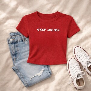 Puede incluir: Una camiseta roja de manga corta y cuello redondo con el texto blanco STAY WEIRD impreso en el pecho, combinada con unos vaqueros de mezclilla azul claro desgastados y un par de zapatillas de lona blancas con cordones y suela de goma.