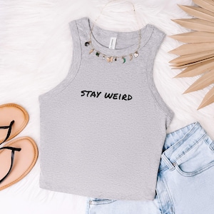 Puede incluir: Un top corto sin mangas de color gris jaspeado con el texto STAY WEIRD impreso en negro en el pecho. El top se combina con un collar dorado con pequeños dijes, pantalones cortos de mezclilla azul claro y sandalias beige con tiras negras.
