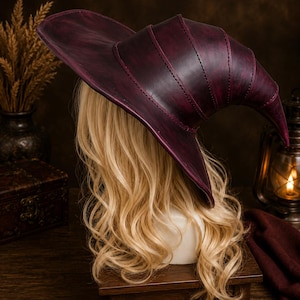 May include: A deep burgundy witch hat constructed from multiple stitched leather panels with a wide, curved brim and a pointed, slightly slumped crown, displayed on a mannequin head with long, wavy blonde hair.