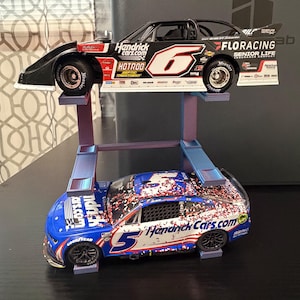 May include: Two die-cast race car models displayed on a two-tier purple stand. The top car is black with the number 6, HendrickCars.com, and Floracing branding. The bottom car is blue and white with the number 5, HendrickCars.com, and confetti graphics.