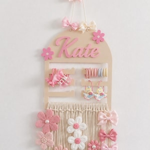 May include: A light wood wall-mounted hair accessory organizer featuring the name Kate in pink script. The top section has slots for hair ties and clips, while the bottom features cream macrame fringe decorated with pink and white felt flowers and bows.