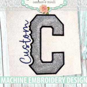 May include: A machine embroidery design featuring a large, silver glitter-textured block letter C with a thick black outline. To the left of the letter, the word Custom is stitched in a dark blue cursive script font on a white fabric background.