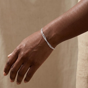 May include: A silver-toned bracelet featuring three thin, intertwined bands that wrap around the wrist. The metallic strands are smooth and reflective, creating a layered appearance that sits securely on the arm with a simple, continuous design.