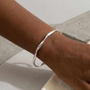 May include: A silver-coloured metal bangle bracelet featuring a continuous, smooth, and undulating wave-like design. The bracelet has a polished, reflective surface that catches the light, creating subtle highlights along its curved, twisted form.