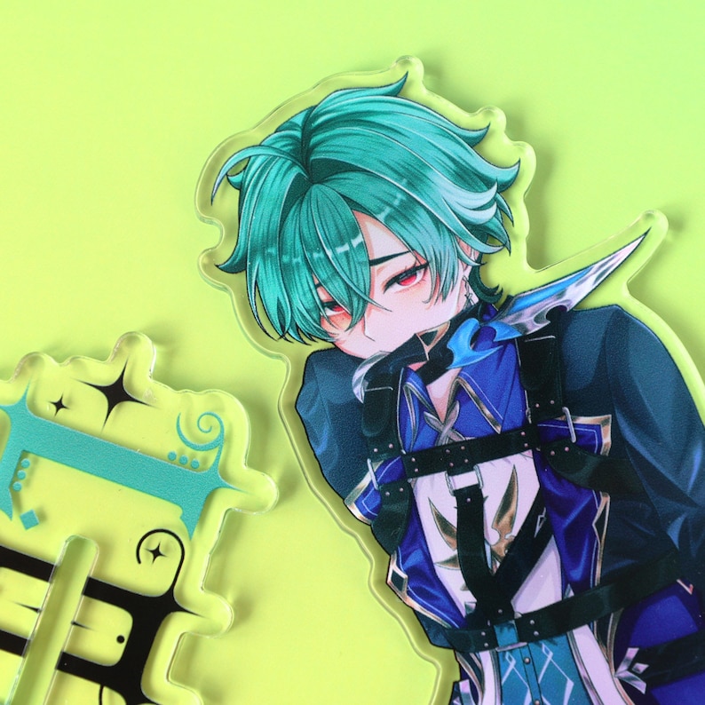 May include: A clear acrylic charm featuring an anime-style character with teal hair and red eyes, wearing a dark blue jacket with black harness straps. Beside it is a separate clear acrylic piece with black and teal decorative line art.