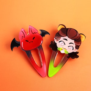 May include: Two hair clips with decorative acrylic toppers. One clip features a red bat with black wings and a gradient red-to-black base. The other clip features a cartoon character with brown hair and yellow ears, set on a green-to-black gradient base.