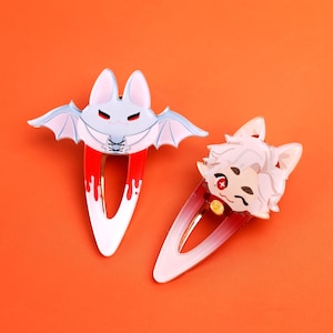 May include: Two metal hair clips featuring enamel designs. One clip depicts a light blue bat character with red accents and a white bodice. The second clip features a light-skinned character with white hair, red eyes, and a pink gradient base.