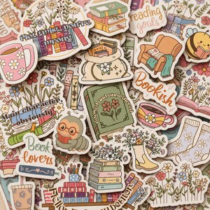 May include: A collection of colourful die-cut stickers featuring book-themed illustrations, including stacks of books, a reading chair, a mug with a smiley face, a caterpillar with glasses, floral arrangements, and text such as Plot twist you're wrong.