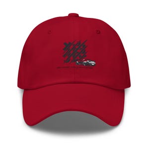 Puede incluir: Una gorra de béisbol roja con un gráfico negro de un coche y caracteres japoneses superpuestos con marcas de arañazos diagonales. Debajo del gráfico, el texto dice JDM OUKAMI / APEX & AXIS CUSTOMS. La gorra tiene una visera curva.