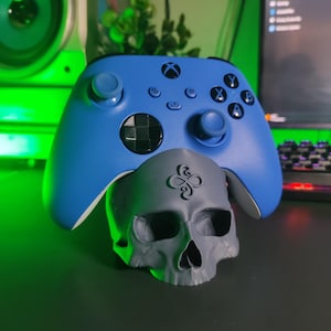 May include: A bright blue wireless video game controller resting on a matte gray skull-shaped stand. The skull features a stylized infinity-like symbol embossed on the forehead area, and the controller has standard black buttons and joysticks.