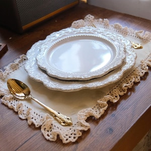 May include: A rectangular cream-colored fabric placemat featuring a decorative crocheted lace border. Centered on the placemat are two stacked white ceramic plates with embossed edges. A polished gold-tone metal spoon rests on the placemat.