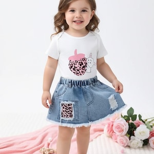 Pode incluir: Um body branco de manga curta com um gráfico de morango rosa com estampa de leopardo e um pequeno coelho branco, combinado com uma saia jeans azul com bainha desfiada, um bolso de remendo com estampa de leopardo e detalhes desgastados.