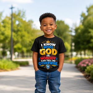 May include: A black short-sleeved t-shirt featuring a graphic print with a gold crown, the text I KNOW WHO GOD IS. NOBODY CAN TELL ME OTHERWISE. in white, yellow, red, and blue, and a blue shield icon with a white cross at the bottom.
