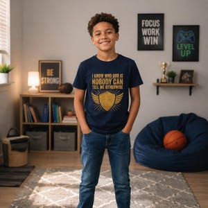 May include: A navy blue short-sleeved t-shirt featuring a gold graphic of a shield with a cross and wings. The text reads: I KNOW WHO GOD IS NOBODY CAN TELL ME OTHERWISE. The shirt is paired with blue denim jeans and white sneakers.