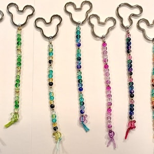 Op de afbeelding: Een collectie van twaalf sleutelhangers, elk voorzien van een zilverkleurige metalen ring in de vorm van een muizenkop-silhouet. Aan elke ring hangt een lange, verticale streng van kleine, veelkleurige doorzichtige kralen met een knoop.