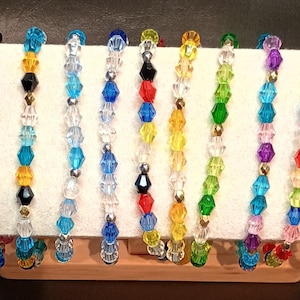 May include: A collection of thirteen beaded bracelets displayed on a horizontal stand. Each bracelet features faceted, multi-coloured crystal beads in various hues including pink, blue, yellow, green, orange, and clear, interspersed with small metallic beads.
