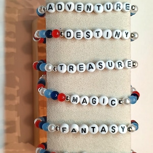 May include: A collection of eight elastic bracelets featuring white letter beads spelling DREAM, ADVENTURE, DESTINY, TREASURED, MAGIC, FANTASY, WONDER, and WISH. The bracelets are accented with round beads in shades of red, light blue, dark blue, and silver.