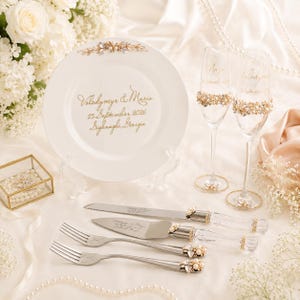 May include: A white ceramic plate with gold text reading Valodymyr & Maria 25 September 2026 Sighnaghi, Georgia, two champagne flutes, a cake knife, and two forks, all featuring gold and pearl embellishments on the handles and stems.