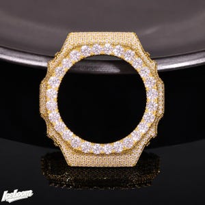 May include: A gold-tone watch bezel featuring an octagonal shape, fully encrusted with numerous small, clear, round-cut stones. The surface is densely paved with these sparkling stones, creating a uniform, reflective, and highly textured appearance.