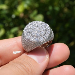 May include: A silver-toned ring featuring a large, circular face densely set with numerous clear, round-cut stones. The band and the sides of the ring are also fully encrusted with smaller clear stones. The text Iceloom is visible on the side.