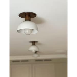 Op de afbeelding: Een plafondlamp met een matwitte, koepelvormige kap en een ronde voet van bronskleurig metaal. De armatuur bevat een enkele, zichtbare gloeilamp in het midden van de kap, die zorgt voor een directe verlichting naar beneden gericht.