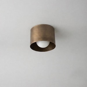 May include: A cylindrical flush mount ceiling light fixture with a brushed antique brass finish. The fixture features a wide, circular metal housing that surrounds a single, centered, spherical white glass light bulb, creating a simple design.