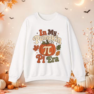 以下が含まれることがあります： 茶色、クリーム色、緑色の文字で「In My Pumpkin Pi Era」と書かれたグラフィックプリントが施された白い長袖スウェットシャツ。中央には数学のパイ記号が描かれたオレンジ色のカボチャがあり、その周りに葉が配置されています。