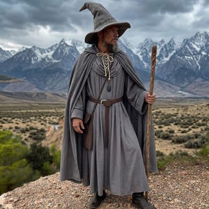 May include: A grey hooded cloak and matching pointed hat worn over a long-sleeved grey tunic with a brown belt and a small pouch. The outfit includes a wooden staff with a twisted texture, featuring a dark pendant hanging from a light-colored cord.