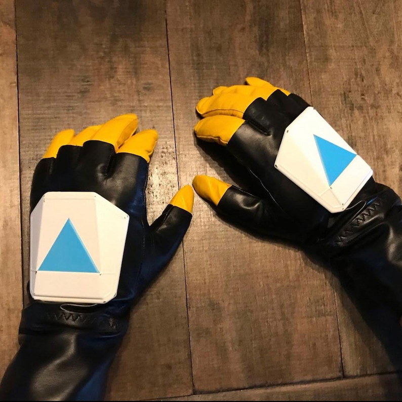 Puede incluir: Un par de guantes negros y amarillos con una placa rectangular blanca en el dorso de cada mano. Cada placa est&aacute; decorada con un tri&aacute;ngulo azul brillante que apunta hacia arriba. Los guantes tienen puntas amarillas y palmas negras.
