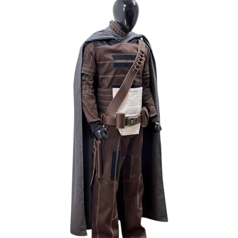 May include: A brown long-sleeved jumpsuit with white chest paneling, a dark gray cape, and a brown utility belt featuring a shoulder strap. The outfit includes visible white stitching details, black accents on the sleeves and legs, and a holster.