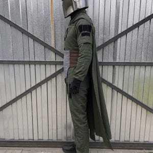 Puede incluir: Un disfraz de cuerpo completo que presenta un casco plateado met&aacute;lico con una visera en forma de T, un mono de manga larga verde oliva, una capa a juego, un cintur&oacute;n marr&oacute;n y gris, guantes negros y zapatillas negras, visto de perfil.