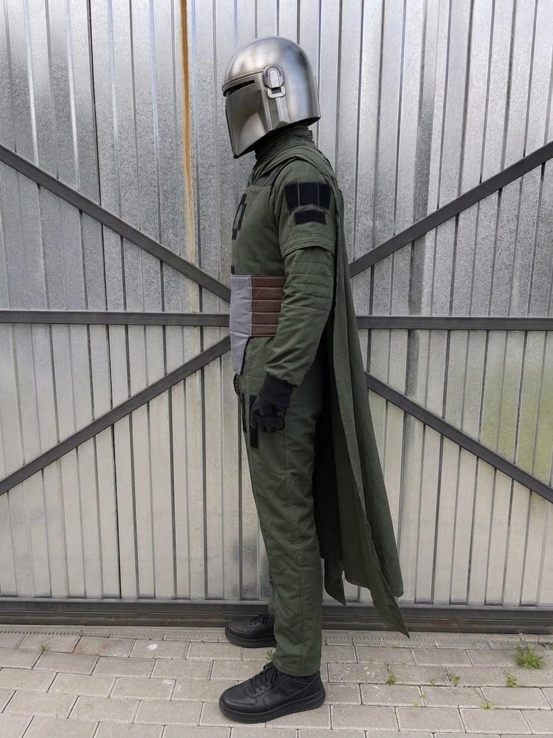 Puede incluir: Un disfraz de cuerpo completo que presenta un casco plateado met&aacute;lico con una visera en forma de T, un mono de manga larga verde oliva, una capa a juego, un cintur&oacute;n marr&oacute;n y gris, guantes negros y zapatillas negras, visto de perfil.