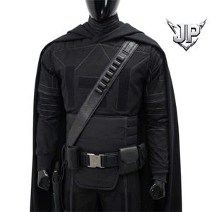 May include: A black tactical costume featuring a long cape, a long-sleeved shirt with geometric stitching, and a utility belt with pouches and a silver buckle. A diagonal shoulder strap with ammunition loops is worn over the chest. The logo JP is visible.