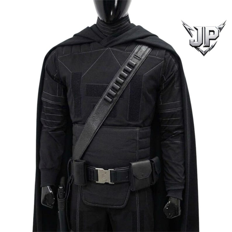 May include: A black tactical costume featuring a long cape, a long-sleeved shirt with geometric stitching, and a utility belt with pouches and a silver buckle. A diagonal shoulder strap with ammunition loops is worn over the chest. The logo JP is visible.