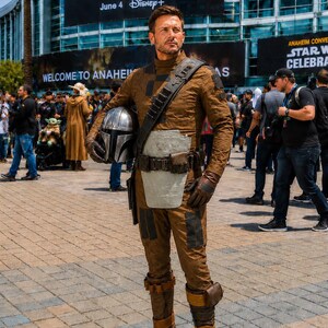 May include: A person wearing a brown textured jumpsuit with a light gray chest plate, a dark brown utility belt, and a shoulder strap. The outfit includes brown boots with straps and dark gloves. The person holds a metallic silver helmet in one hand.