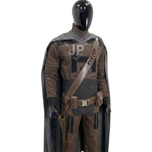 May include: A brown and black tactical costume set on a mannequin, featuring a long grey cape, a high-neck long-sleeve top, matching trousers, and a brown utility belt with a holster and pouches. The chest area displays the letters JP in grey.