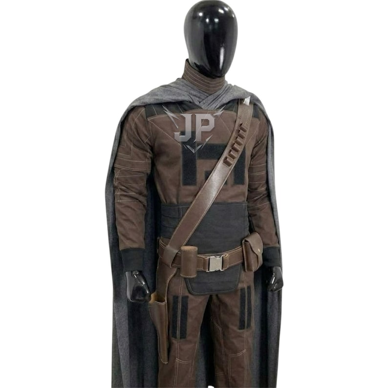 May include: A brown and black tactical costume set on a mannequin, featuring a long grey cape, a high-neck long-sleeve top, matching trousers, and a brown utility belt with a holster and pouches. The chest area displays the letters JP in grey.