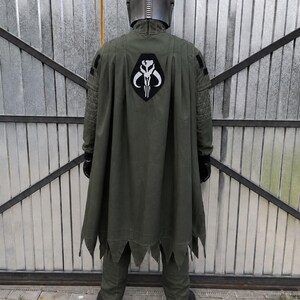 Puede incluir: Un disfraz de cuerpo completo que incluye un casco de color plata met&aacute;lica y una chaqueta verde oliva con mangas acolchadas. Una capa larga a juego de color verde oliva cuelga de los hombros, mostrando un emblema de cr&aacute;neo de Mythosaurio en la espalda.