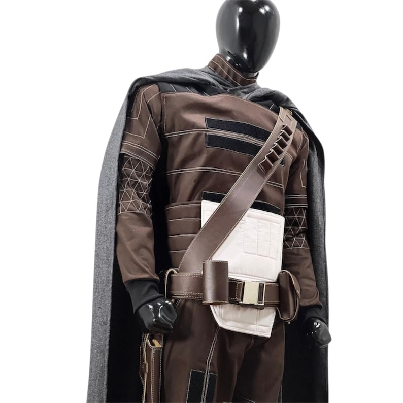 May include: A brown long-sleeved costume set featuring a dark grey cape, a white chest panel, and a brown utility belt with pouches. The outfit includes intricate white stitching details on the sleeves and torso, with a bandolier strap across the chest.