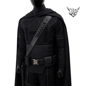 May include: A black costume set featuring a long cape, a long-sleeved shirt with subtle geometric stitching, and a utility belt with multiple pouches and a silver buckle. A diagonal shoulder strap with ammunition loops crosses the chest.