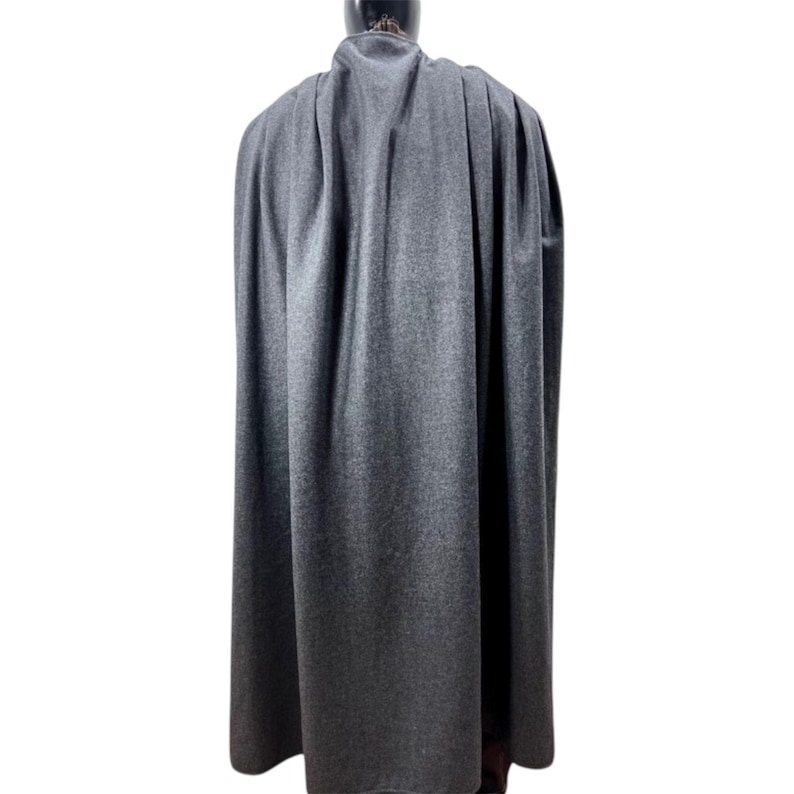 May include: A floor-length, dark charcoal gray cape featuring a textured, wool-like fabric. The garment is draped over a mannequin, displaying vertical folds and pleats that extend from the high, structured collar down to the hemline.