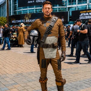 May include: A person wearing a brown textured long-sleeve shirt and matching trousers with dark knee patches. The outfit includes a grey chest plate, a dark utility belt with a shoulder strap, and brown boots. A silver helmet is held under one arm.