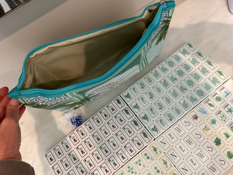 May include: A teal zipped pouch with a botanical print containing a set of mahjong tiles. The tiles feature various green and blue illustrations, Chinese characters, and the word NEWS. A small plastic bag with blue dice is included in the set.