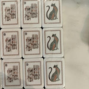 May include: A set of rectangular white mahjong tiles arranged in a grid. The tiles feature printed designs including the number 8 with bamboo stalks, the number 9 with bamboo stalks, and a stylized illustration of a seated cat in gray and pink.