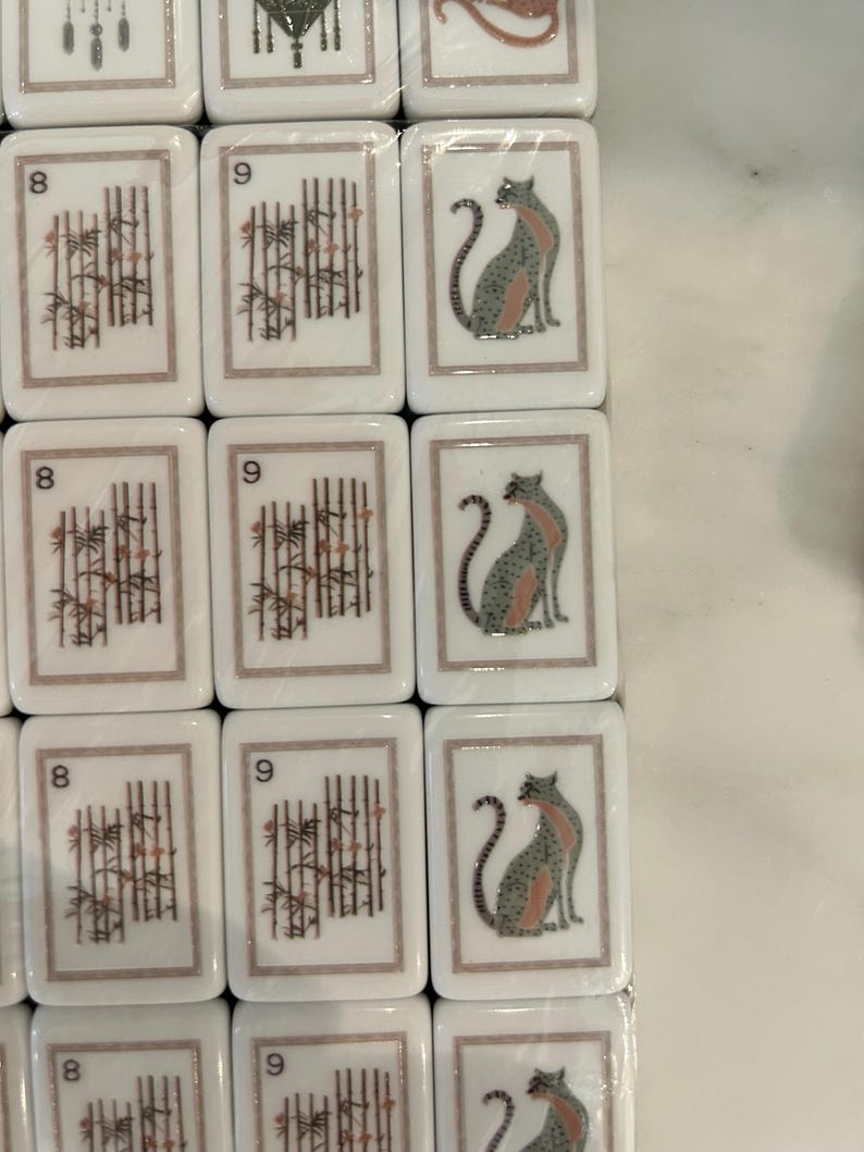 May include: A set of rectangular white mahjong tiles arranged in a grid. The tiles feature printed designs including the number 8 with bamboo stalks, the number 9 with bamboo stalks, and a stylized illustration of a seated cat in gray and pink.