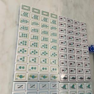 May include: A collection of white Mahjong tiles arranged in a grid. The tiles feature various green floral patterns, Chinese characters, and the word NEW printed in blue. Some tiles display numerical values, while others show intricate green designs.
